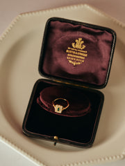 The CAMELLIA Seraphina Emerald Ring, featuring an emerald-green center, is showcased in a maroon velvet box with gold lettering on the lid atop a white plate, radiating timeless heirloom elegance.
