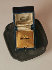 The CAMELLIA Elowen Peridot Ring, featuring a heart-cut peridot in 14K gold vermeil, comes in a blue velvet box and is elegantly displayed on a dark ceramic dish.