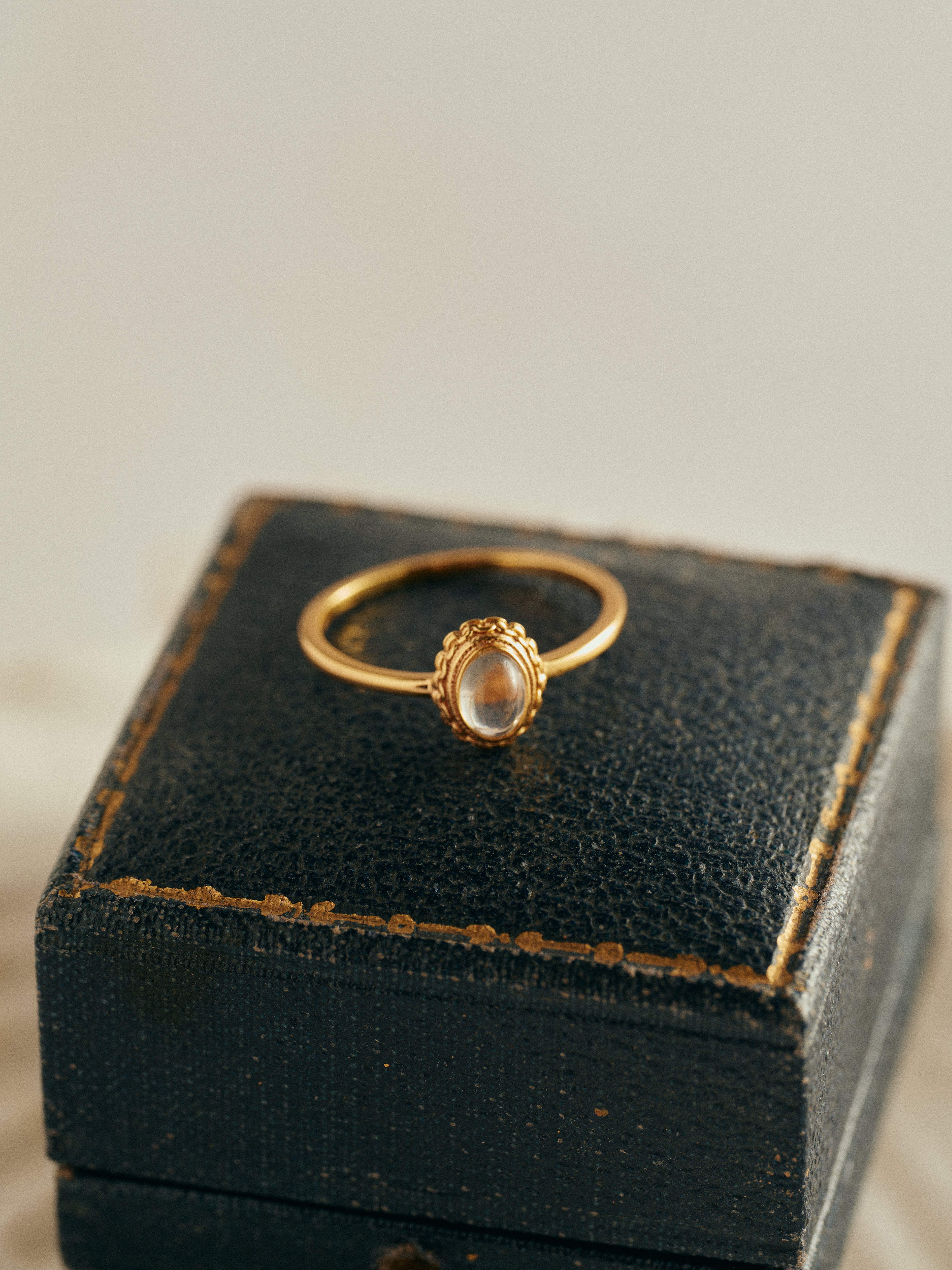 The CAMELLIA Eloise Moonstone Ring, a vintage-inspired piece with an oval-cut moonstone, is displayed on a black, textured jewelry box with worn edges.