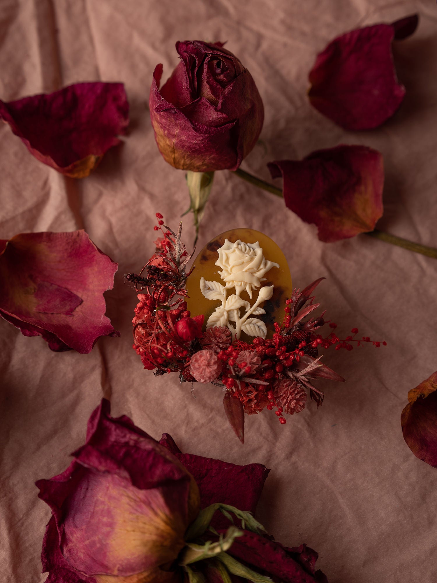 Red roses and petals are artfully arranged with a CAMELLIA Rose Birth Flower Ring, handcrafted in New York, set on a beige fabric background.