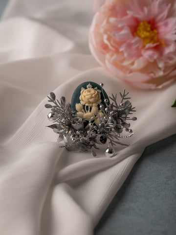 A handcrafted decorative brooch with a white rose on dark green, framed by silver floral designs, sits on white fabric. In the background, a pink peony echoes the elegance of our CAMELLIA Cotton Rose Hibiscus Birth Flower Ring collection.
