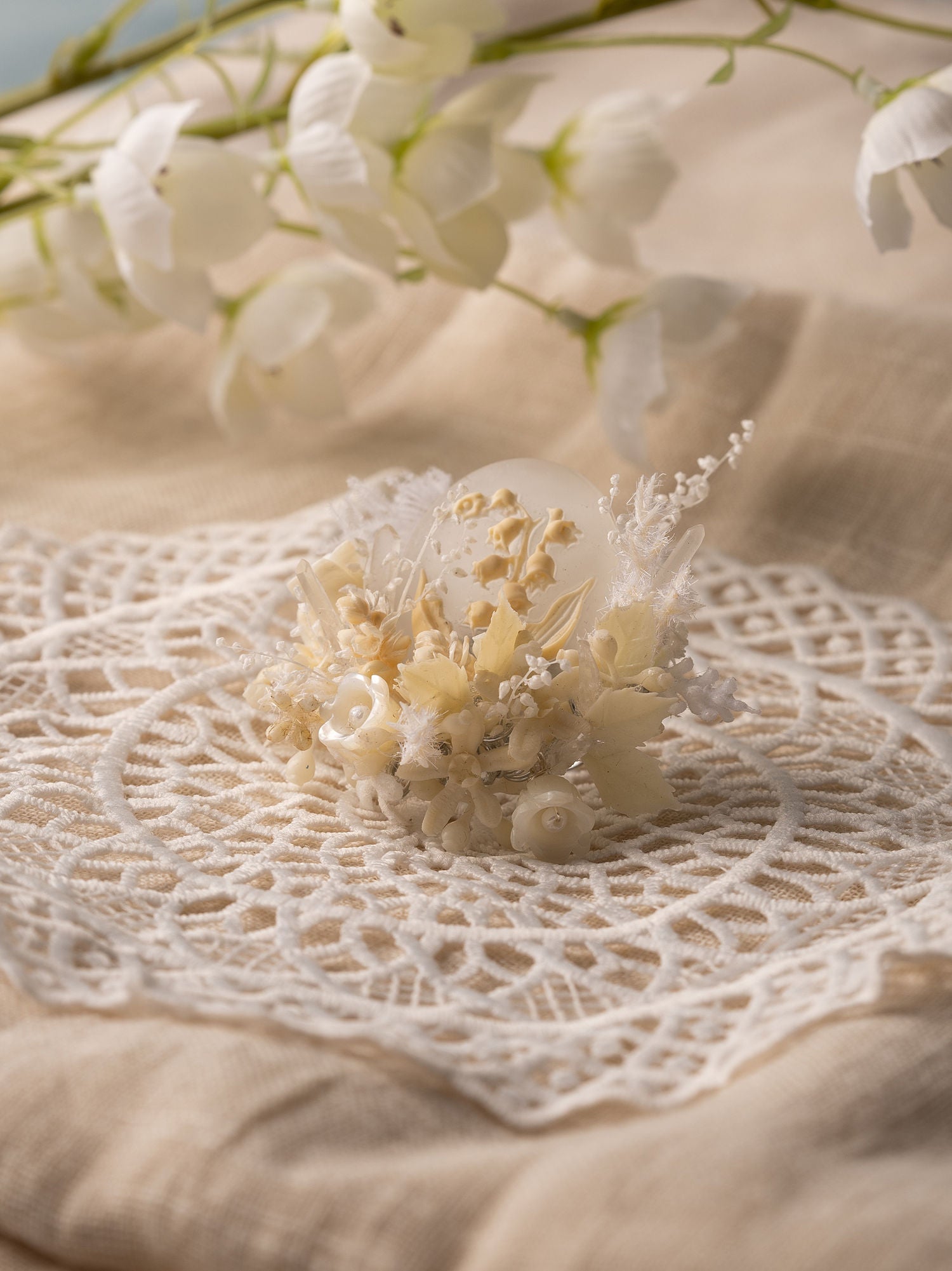 A small, delicate floral arrangement with white and cream flowers sits on an intricate crocheted doily, reflecting the elegance of CAMELLIA's Lily of the Valley Birth Flower Ring handcrafted in New York, as blurred white flowers linger in the background.