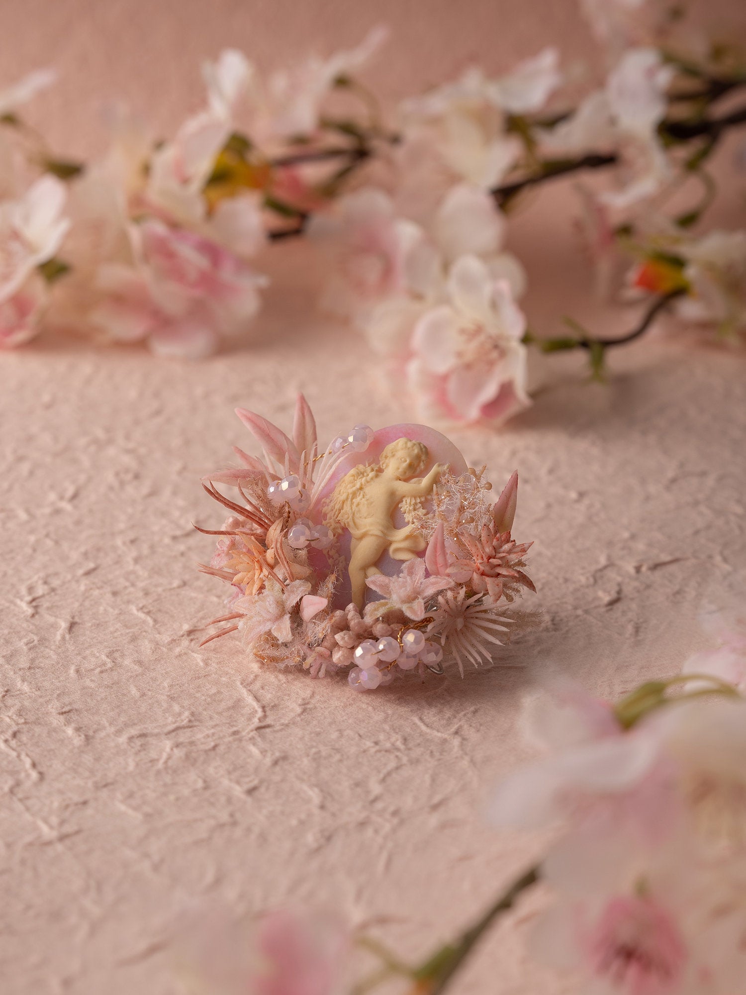 The CAMELLIA Cherry Blossom Birth Flower Ring is an exquisite handcrafted piece from New York, featuring a small figure amidst pink and white flowers on a textured surface. The blurred blossoms in the background add depth and charm.