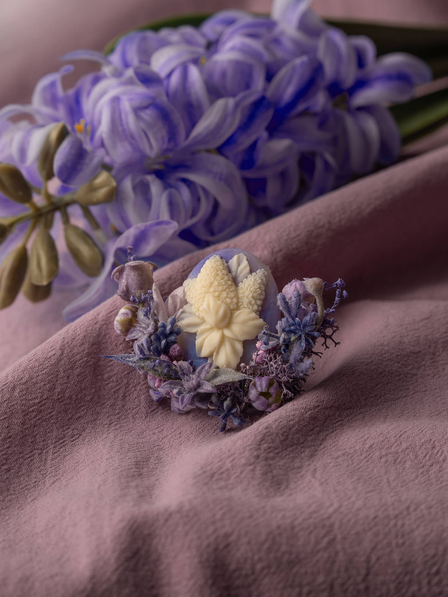 The CAMELLIA Hyacinthus Birth Flower Ring is delicately crafted in New York and features a detailed white floral wax seal accented by purple and blue dried flowers on textured pink fabric, with a graceful touch of purple hyacinth.