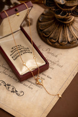 The CAMELLIA Aurielle Opal Long Necklace gleams elegantly on an open jewelry box, resting on vintage-style paper with decorative text, beside a bronze candle holder.