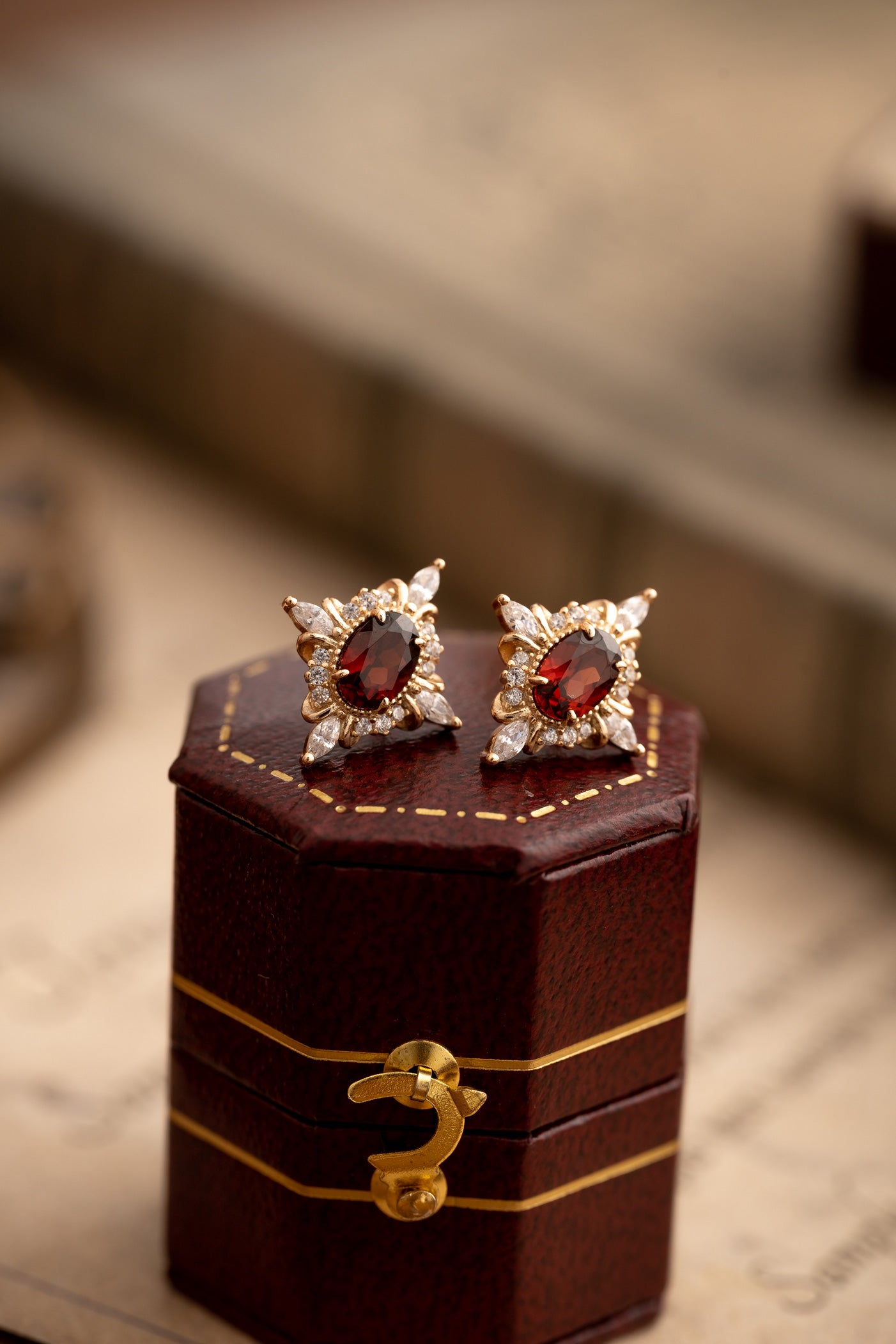 The Claire Garnet Diamond Jewelry Set by CAMELLIA features garnet stones in gold star-shaped earrings with clear gems, presented in a hexagonal brown box with a golden latch. Hypoallergenic and stunning, these pieces add elegance to any collection against a softly blurred backdrop.