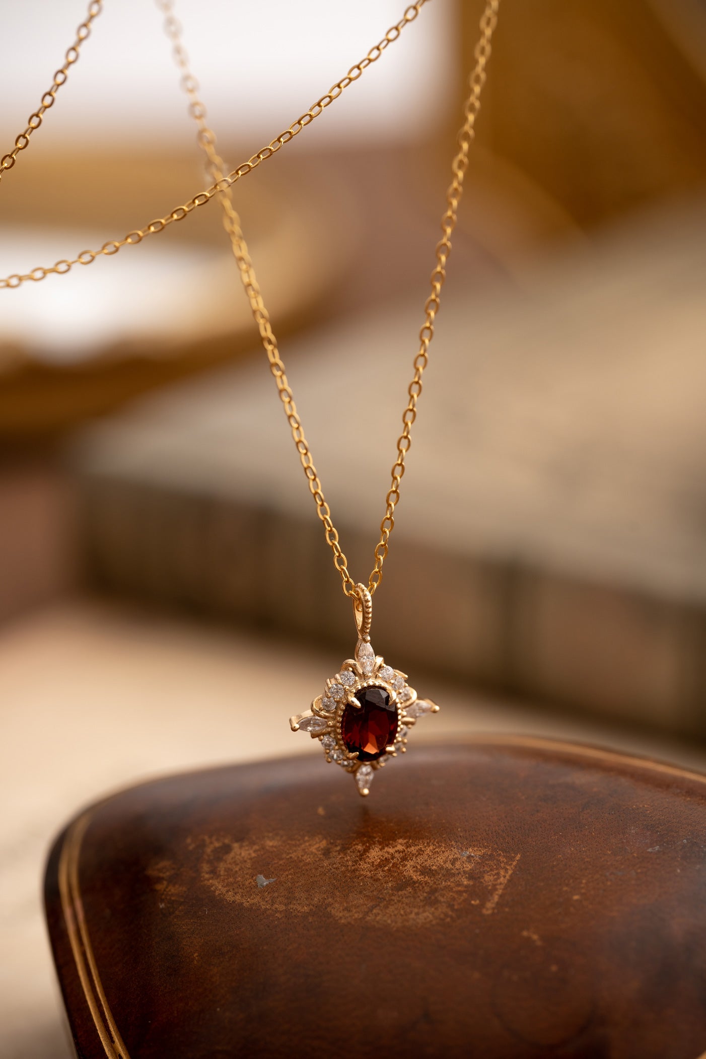 The Claire Garnet Diamond Jewelry Set by CAMELLIA features a stunning gold necklace with a garnet gemstone pendant and small diamonds, elegantly displayed on brown leather.