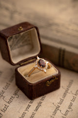 The Claire Garnet Diamond Jewelry Set by CAMELLIA features a stunning garnet ring with a dark center stone, surrounded by smaller gems, crafted from hypoallergenic sterling silver. It graces a vintage brown box resting on paper adorned with faded cursive writing.