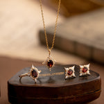 The Claire Garnet Diamond Jewelry Set by CAMELLIA, featuring a necklace, ring, and earrings adorned with vibrant garnet gemstones, is elegantly displayed on a brown leather box.