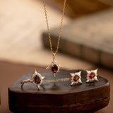 The Claire Garnet Diamond Jewelry Set by CAMELLIA, featuring a necklace, ring, and earrings adorned with vibrant garnet gemstones, is elegantly displayed on a brown leather box.