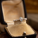 The CAMELLIA Starlight Opal Jewelry Set features a handcrafted teardrop-shaped opal ring with diamond accents set in sterling silver, elegantly displayed in a vintage velvet-lined box on a wooden surface.