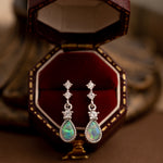 A pair of handcrafted teardrop-shaped opal and diamond earrings from the Starlight Opal Jewelry Set by CAMELLIA rests in an open red velvet box, set against a soft-focus background.