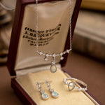 The CAMELLIA Starlight Opal Jewelry Set includes a handcrafted necklace, earrings, and ring in an open brown box. These Sterling Silver pieces feature exquisite opal gemstones with diamond-like accents.
