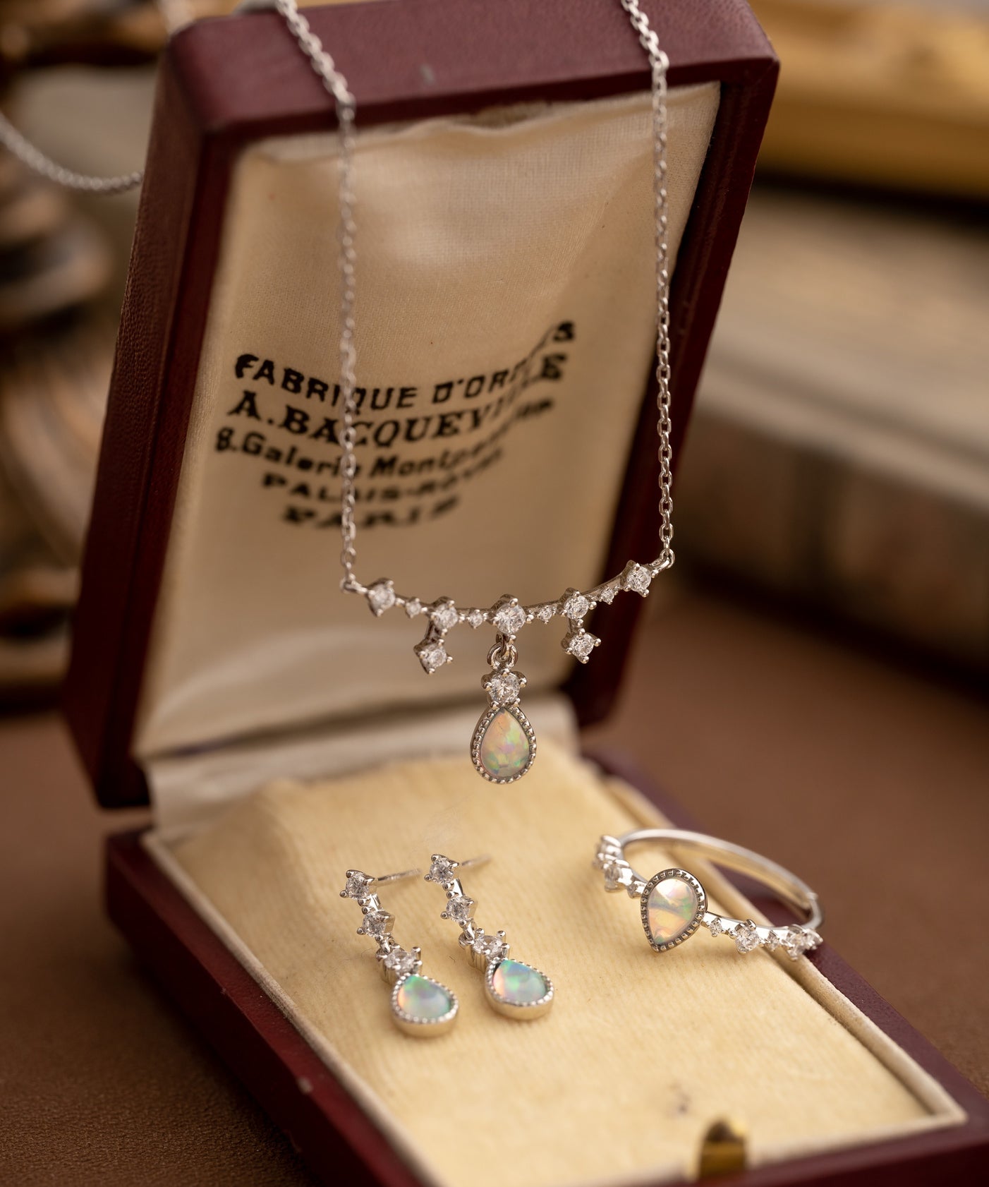 The CAMELLIA Starlight Opal Jewelry Set includes a handcrafted necklace, earrings, and ring in an open brown box. These Sterling Silver pieces feature exquisite opal gemstones with diamond-like accents.