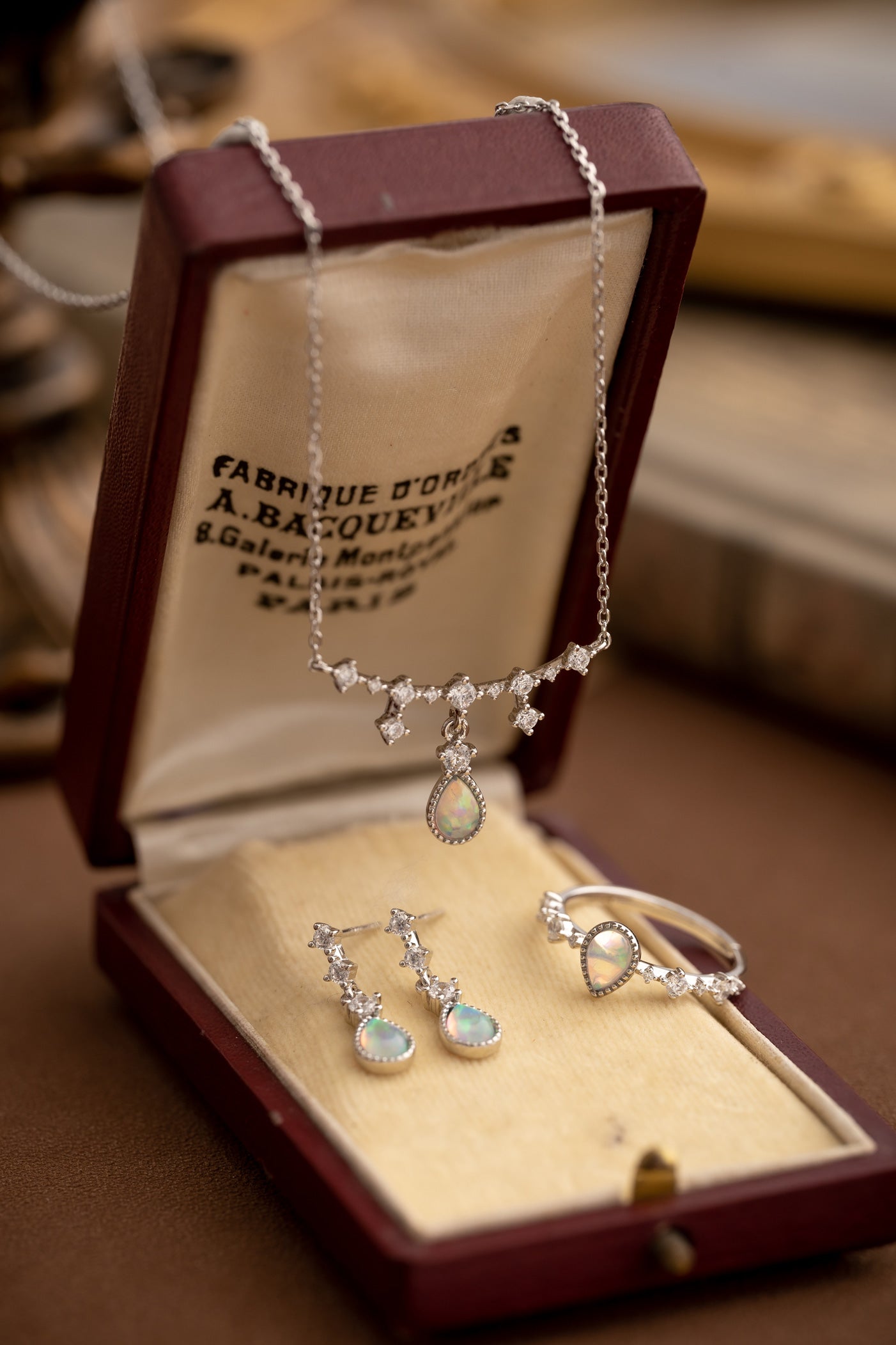 The CAMELLIA Starlight Opal Jewelry Set includes a handcrafted necklace, earrings, and ring in an open brown box. These Sterling Silver pieces feature exquisite opal gemstones with diamond-like accents.