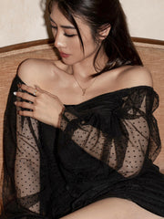 A woman in a sheer black, off-shoulder polka-dot dress sits on a couch, looking down and touching her shoulder, as the CAMELLIA Evermine Moissanite Band with pavé-set accents sparkles in the light.