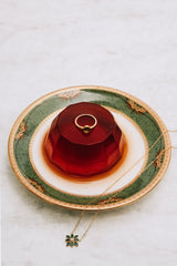 The CAMELLIA Eden Peridot Ring with a vintage-inspired green stone is displayed atop a red gelatin dessert on a decorative plate; a gold necklace with a matching green pendant lies next to the plate.
