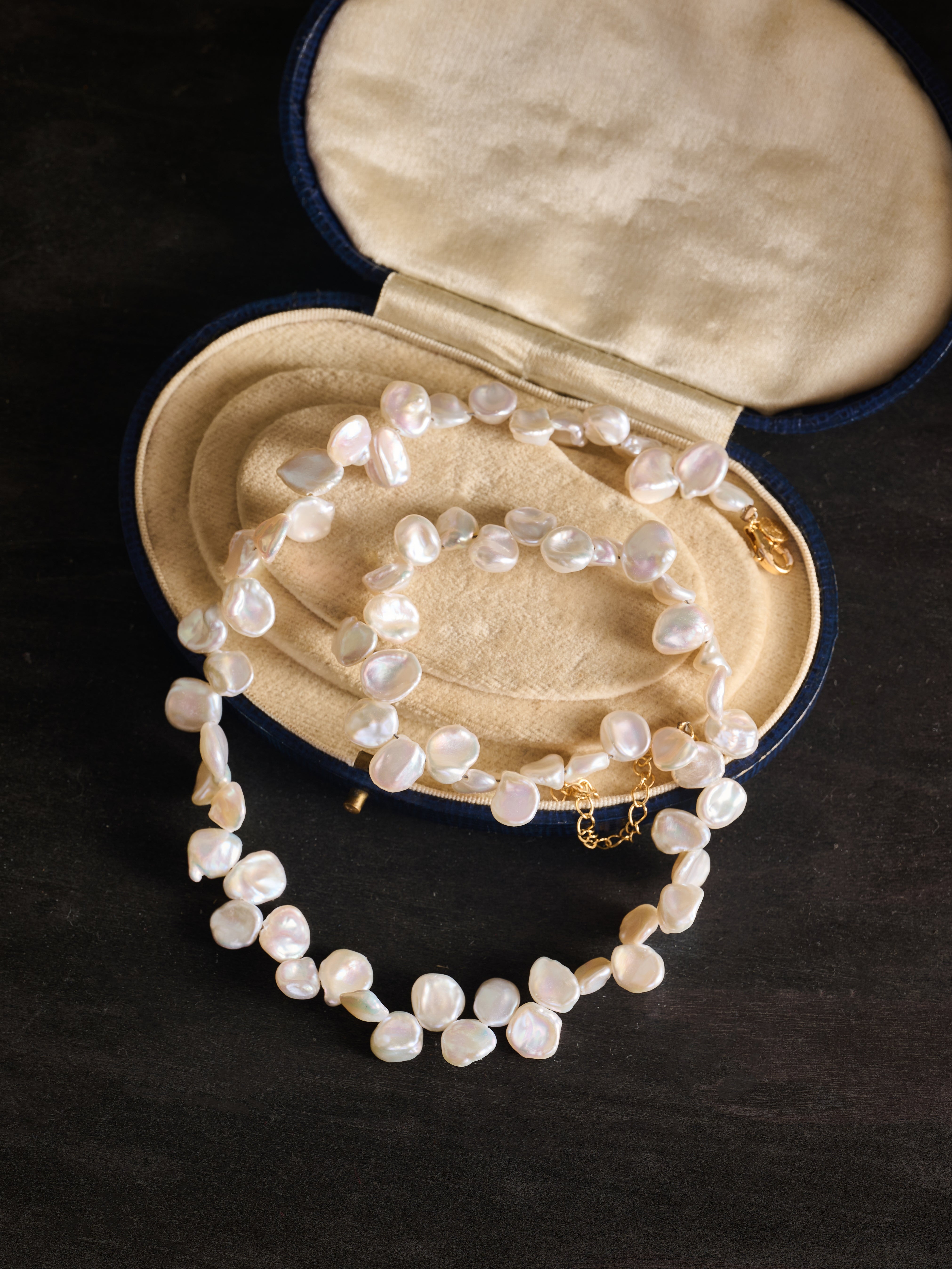 The CAMELLIA Petal Pearl Necklace, a baroque-style freshwater pearl piece, is showcased in an open cream-lined oval jewelry case on a dark surface.