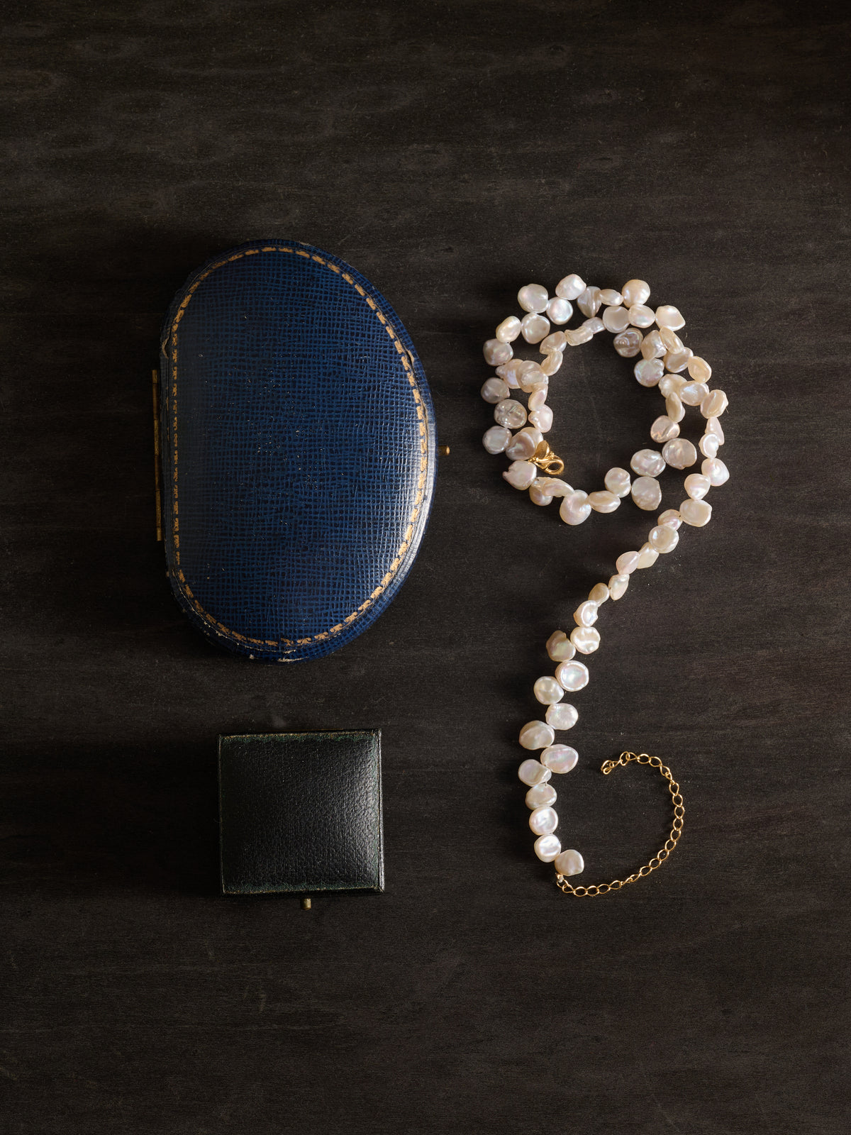 A CAMELLIA Petal Pearl Necklace is arranged in a question mark shape alongside a blue oval jewelry box and a black square box on a dark surface.