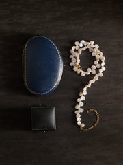 A CAMELLIA Petal Pearl Necklace is arranged in a question mark shape alongside a blue oval jewelry box and a black square box on a dark surface.