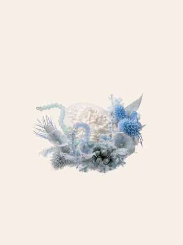 The Winteersweet Birth Flower Ring by CAMELLIA showcases a delicate arrangement of dried preserved white flowers and blue accents with beads and textured foliage on a simple beige background.