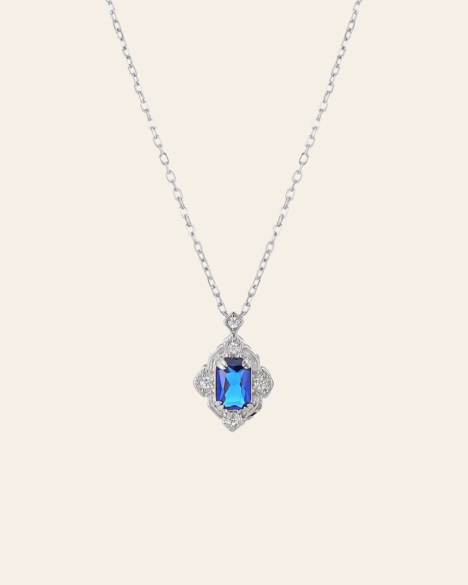 The Louisa Sapphire Necklace by CAMELLIA features a sterling silver chain and a square blue sapphire gemstone pendant surrounded by small clear stones.