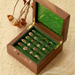 An open wooden box reveals a collection of ornate CAMELLIA rings, including the stunning Mystery Gemstone Ring in yellow gold and sterling silver, all displayed on a green velvet lining resting on a textured surface with dried flowers and lace for an elegant scene.