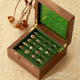 An open wooden box reveals a collection of ornate CAMELLIA rings, including the stunning Mystery Gemstone Ring in yellow gold and sterling silver, all displayed on a green velvet lining resting on a textured surface with dried flowers and lace for an elegant scene.