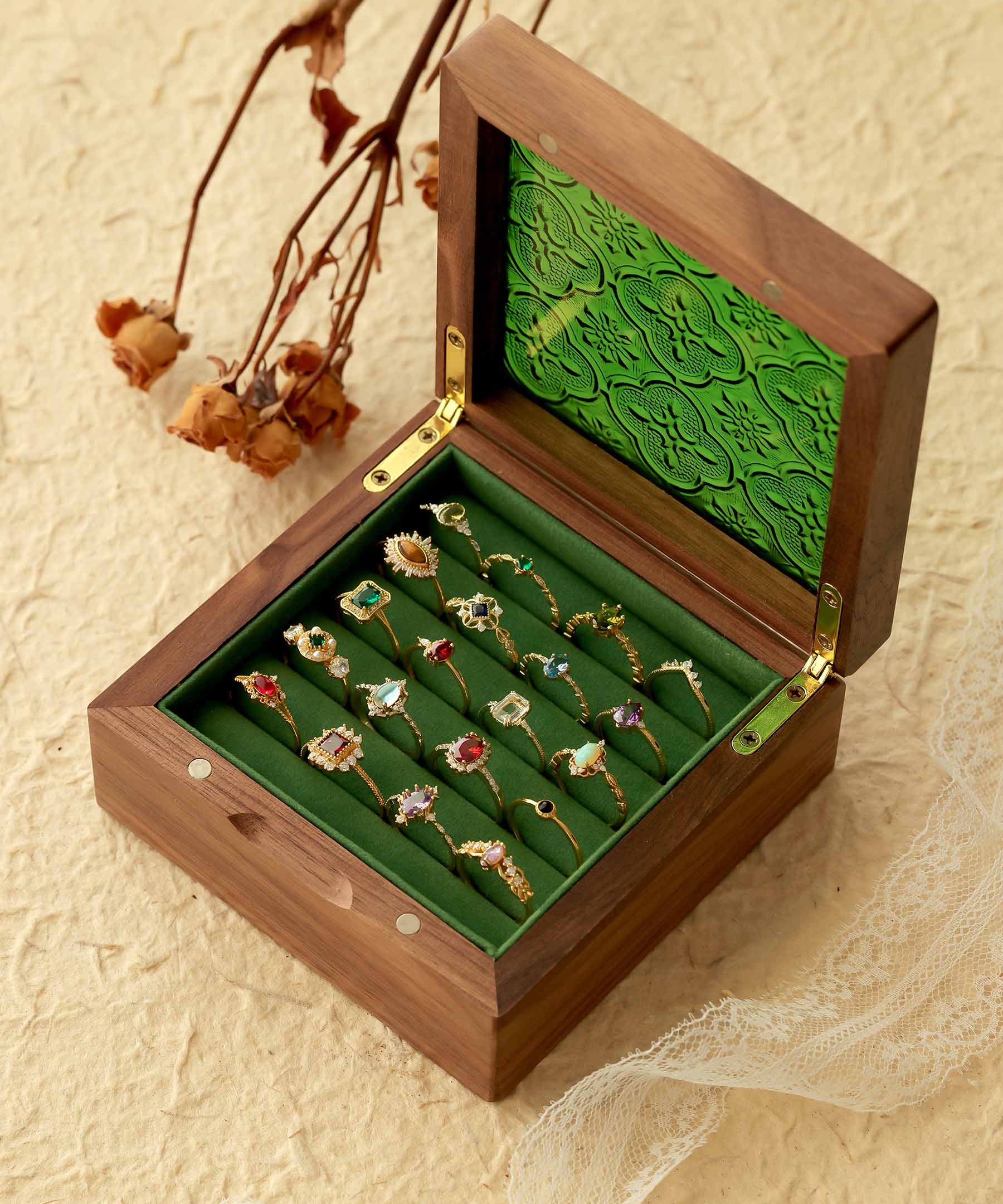 An open wooden box reveals a collection of ornate CAMELLIA rings, including the stunning Mystery Gemstone Ring in yellow gold and sterling silver, all displayed on a green velvet lining resting on a textured surface with dried flowers and lace for an elegant scene.