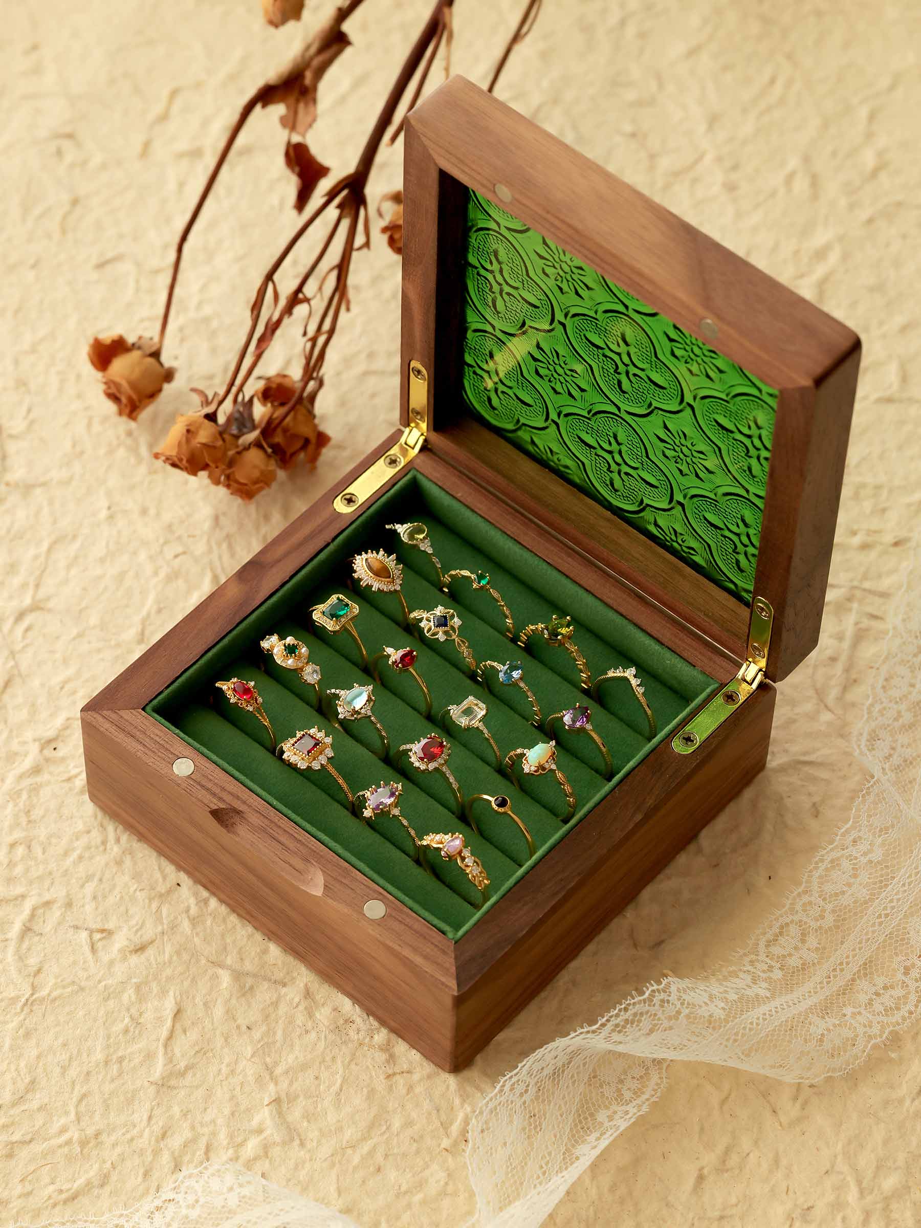 An open wooden box reveals a collection of ornate CAMELLIA rings, including the stunning Mystery Gemstone Ring in yellow gold and sterling silver, all displayed on a green velvet lining resting on a textured surface with dried flowers and lace for an elegant scene.
