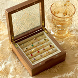 A wooden box with a glass lid displays CAMELLIA's Mystery Gemstone Rings on a lace tablecloth, next to a transparent glass cup of light-colored liquid. Glimpses of yellow gold and sterling silver add elegance to the assorted colorful rings.