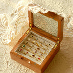 An open wooden jewelry box showcasing CAMELLIA's Mystery Gemstone Rings in yellow gold with assorted colored gemstones is set on a textured surface adorned with lace fabric.