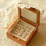 An open wooden jewelry box showcasing CAMELLIA's Mystery Gemstone Rings in yellow gold with assorted colored gemstones is set on a textured surface adorned with lace fabric.