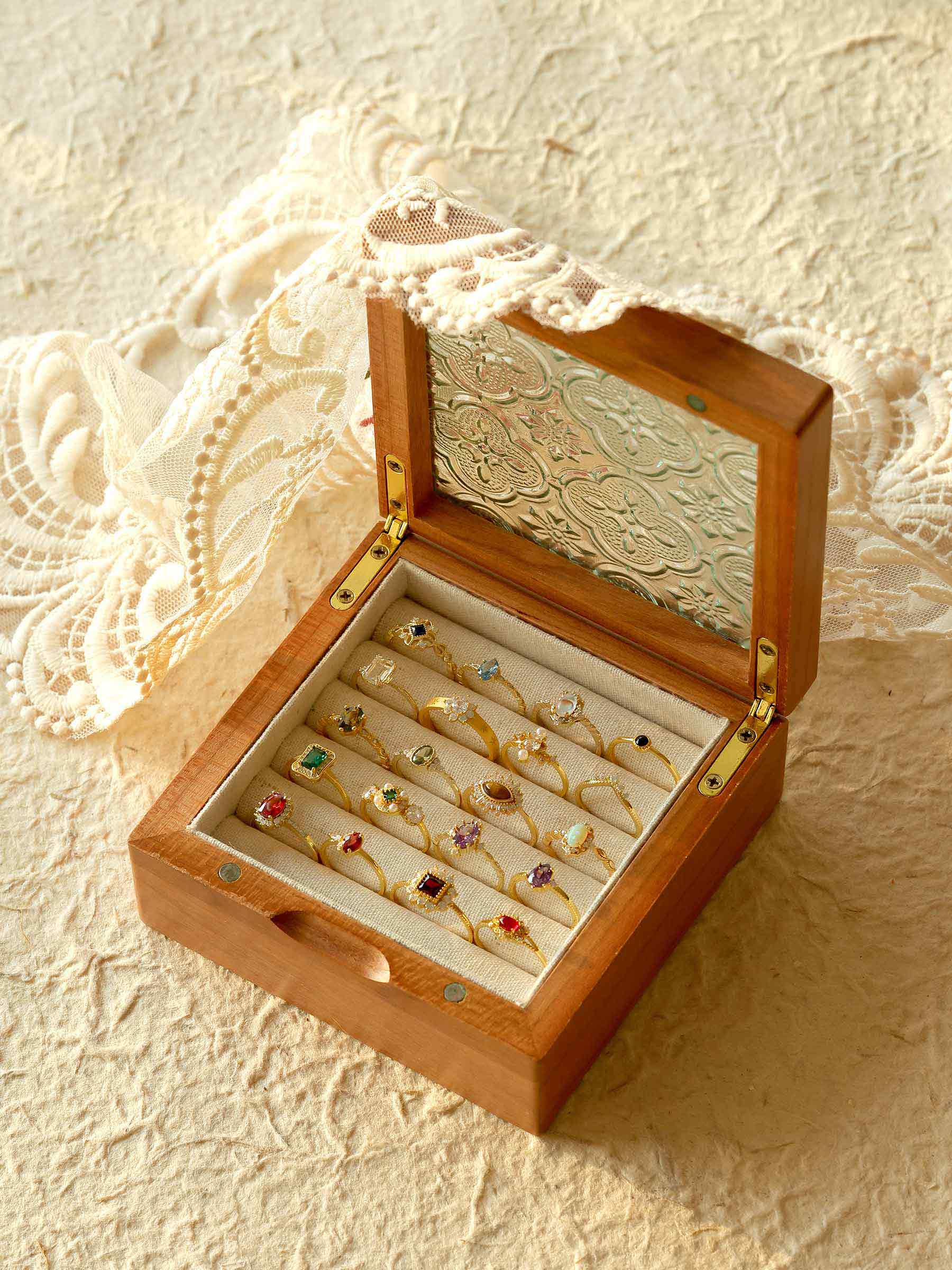 An open wooden jewelry box showcasing CAMELLIA's Mystery Gemstone Rings in yellow gold with assorted colored gemstones is set on a textured surface adorned with lace fabric.
