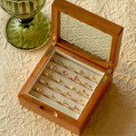 A CAMELLIA Mystery Gemstone Ring rests in a wooden box with an ornate glass lid among vibrant rings, some in sterling silver. A green glass goblet is nearby on a textured surface.