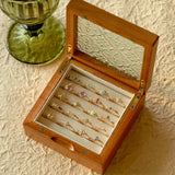 A CAMELLIA Mystery Gemstone Ring rests in a wooden box with an ornate glass lid among vibrant rings, some in sterling silver. A green glass goblet is nearby on a textured surface.