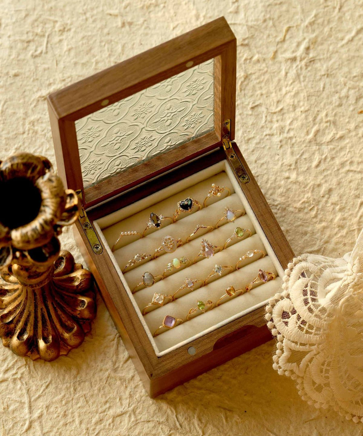 A wooden jewelry box with a glass lid showcases an assortment of jewelry, including the CAMELLIA Mystery Gemstone Ring in sterling silver. Nearby, a vintage candlestick holder and lace fabric rest on the textured surface, completing an elegant display.