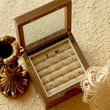 A wooden jewelry box with a glass lid showcases an assortment of jewelry, including the CAMELLIA Mystery Gemstone Ring in sterling silver. Nearby, a vintage candlestick holder and lace fabric rest on the textured surface, completing an elegant display.