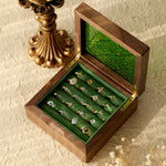 A wooden box with a green interior features CAMELLIA's ornate Mystery Gemstone Rings, including yellow gold pieces. Nearby, a decorative bronze candlestick rests on a textured surface amid dried flowers.