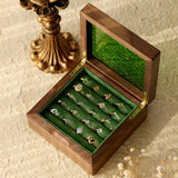 A wooden box with a green interior features CAMELLIA's ornate Mystery Gemstone Rings, including yellow gold pieces. Nearby, a decorative bronze candlestick rests on a textured surface amid dried flowers.