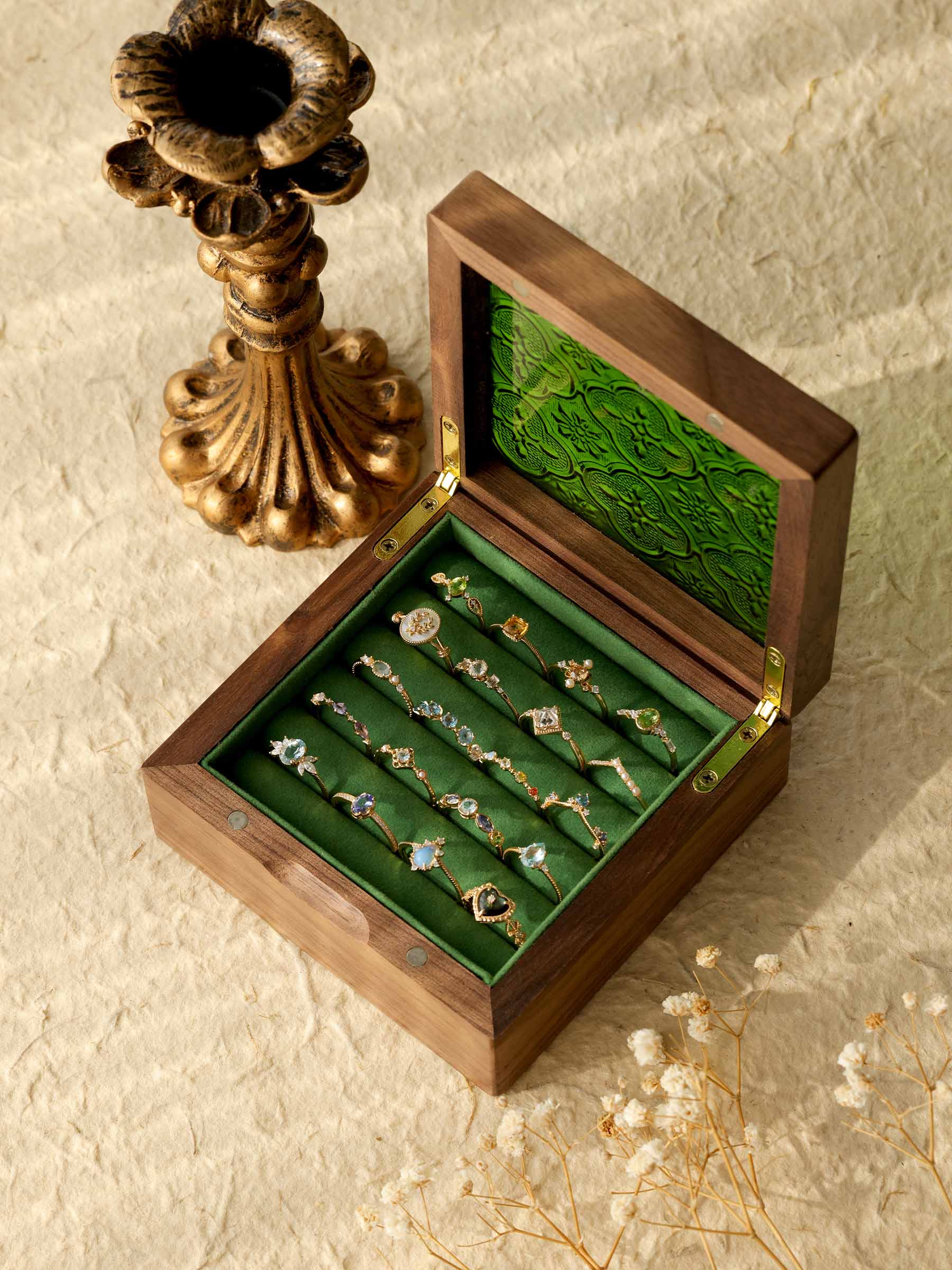 A wooden box with a green interior features CAMELLIA's ornate Mystery Gemstone Rings, including yellow gold pieces. Nearby, a decorative bronze candlestick rests on a textured surface amid dried flowers.