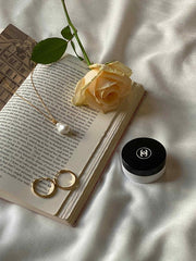 An open book with a yellow rose is beside a jar with a logo, while the Alyssa Barquoe Pearl Necklace by CAMELLIA elegantly drapes across two 14K gold vermeil rings on the white fabric surface.
